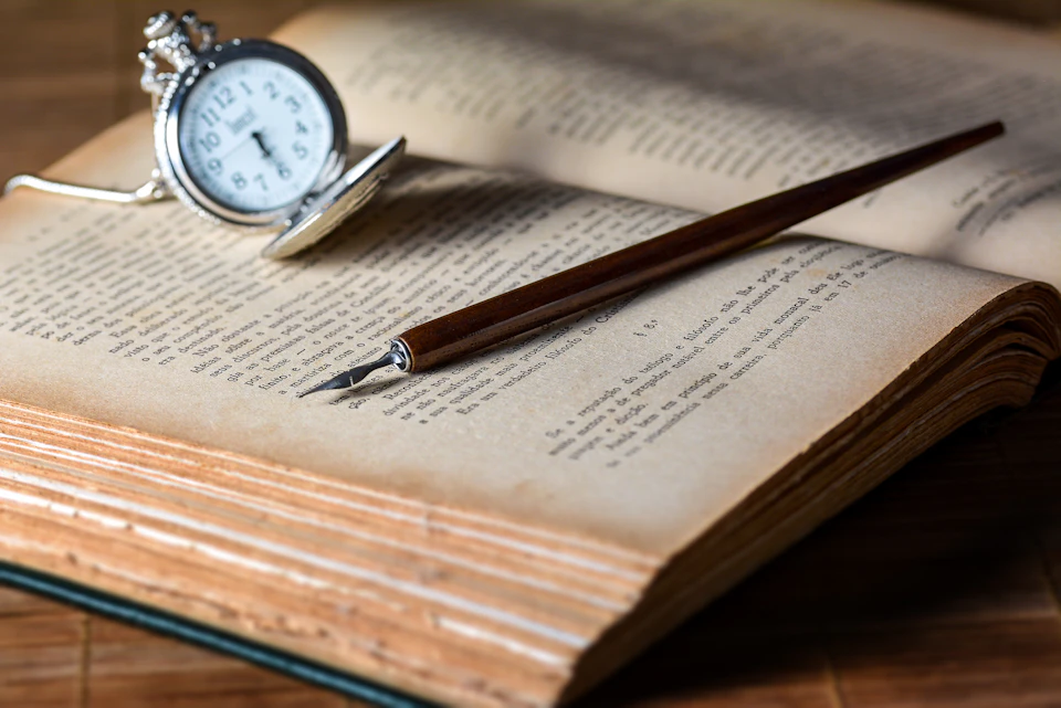 Open book rests on wooden table with brown pen and silver pocket watch nearby