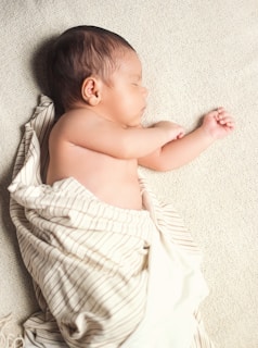 A newborn baby is peacefully sleeping on a soft, textured blanket. The baby is wrapped in a light-colored blanket with thin stripes, leaving one arm exposed. The infant's skin tone is warm and smooth, and their hair is dark.