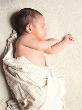A newborn baby is peacefully sleeping on a soft, textured blanket. The baby is wrapped in a light-colored blanket with thin stripes, leaving one arm exposed. The infant's skin tone is warm and smooth, and their hair is dark.