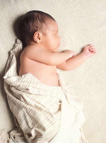 A newborn baby is peacefully sleeping on a soft, textured blanket. The baby is wrapped in a light-colored blanket with thin stripes, leaving one arm exposed. The infant's skin tone is warm and smooth, and their hair is dark.