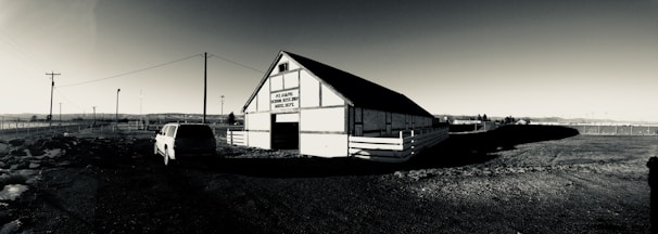 A black and white image features a barn structure with a sign on it reading 'Pvt. ID. Barnes, Horse Det., CO. F., Absentee Depot'. The barn is positioned at an angle, casting a shadow to the right. A vehicle is parked in front of the barn, and there are utility poles and fencing extending into the background. The landscape is flat with distant structures on the horizon.