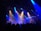 A live band is performing on a dimly lit stage with vibrant blue and orange spotlights illuminating the scene. The audience is visible in the foreground, casting silhouettes against the bright stage lights. Various instruments, including guitars and drums, are seen being played by the musicians.