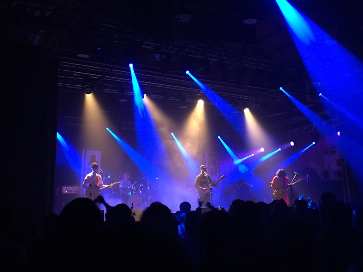 A live band is performing on a dimly lit stage with vibrant blue and orange spotlights illuminating the scene. The audience is visible in the foreground, casting silhouettes against the bright stage lights. Various instruments, including guitars and drums, are seen being played by the musicians.