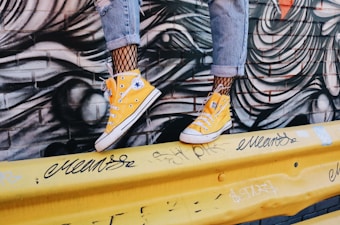 A pair of yellow high-top sneakers is worn with fishnet socks and rolled-up denim jeans. The scene is set against a graffiti-covered brick wall and a yellow barricade adorned with black tags and scribbles.