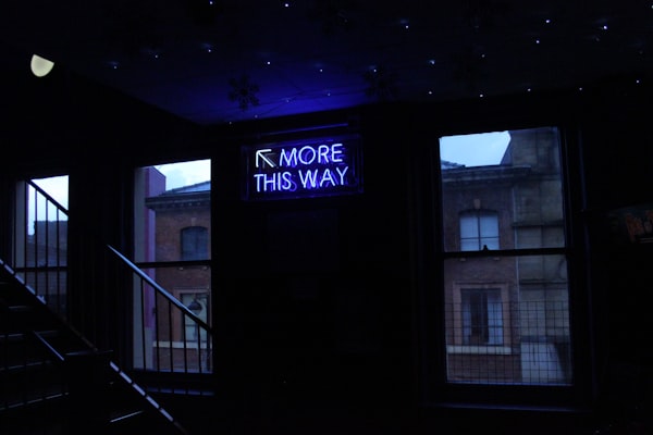 a neon sign, reading "more this way," with a arrow pointing up and to left, by a staircase, in a dark room 