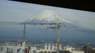 A snow-capped mountain, likely Mount Fuji, stands majestically in the background under a cloudy sky. In the foreground, industrial buildings, electrical poles, and wires crisscross the view, adding an urban element to the natural landscape.