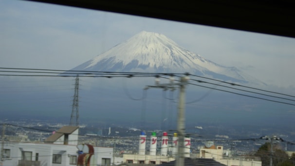 A snow-capped mountain, likely Mount Fuji, stands majestically in the background under a cloudy sky. In the foreground, industrial buildings, electrical poles, and wires crisscross the view, adding an urban element to the natural landscape.