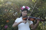 A girl with a floral crown plays the violin in a verdant outdoor setting. Sunlight filters through the trees, casting a serene and natural ambiance.