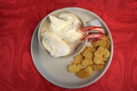 A white mug filled with a hot chocolate topped with whipped cream is resting on a white plate. The mug has a candy cane hanging on the handle. Next to the mug on the plate are several small, light-colored cookies. The background features a red, swirling pattern.