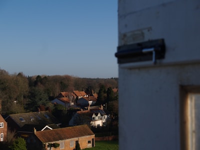 A view of a rural area with several houses featuring red-tiled roofs surrounded by trees. The foreground includes part of a white structure with a visible latch, while the background has a clear blue sky.
