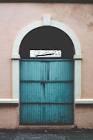 A vintage-style wooden door painted in turquoise with a semicircular transom at the top. The door features vertical panels and is surrounded by a soft pink, slightly worn-out wall.