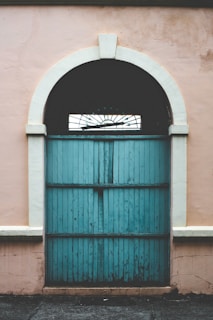 A vintage-style wooden door painted in turquoise with a semicircular transom at the top. The door features vertical panels and is surrounded by a soft pink, slightly worn-out wall.