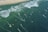 Aerial view of a beach with multiple kite surfers riding the waves. The shoreline is dotted with people on the sand and beach umbrellas. The ocean is a deep green with white crests from the waves, and colorful kites are visible across the water.