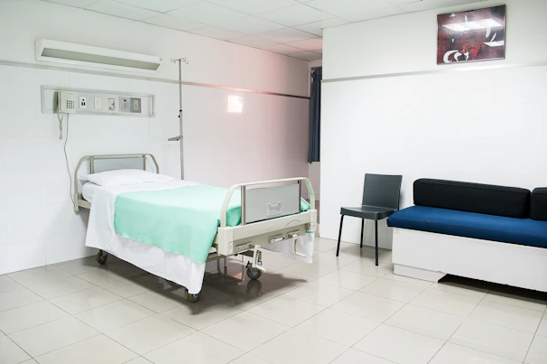 A clean and minimal hospital room with a single hospital bed featuring a white sheet and a green blanket. The room also has medical equipment attached to the wall above the bed, a black chair, and a blue cushioned bench. The walls and floor are white, and there is a soft, fluorescent lighting.