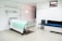 A clean and minimal hospital room with a single hospital bed featuring a white sheet and a green blanket. The room also has medical equipment attached to the wall above the bed, a black chair, and a blue cushioned bench. The walls and floor are white, and there is a soft, fluorescent lighting.