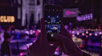 Hands holding a smartphone capturing a dimly lit club or event setting, with low lighting and neon lights creating a vibrant and atmospheric background. The smartphone's screen shows a live view of the surroundings.