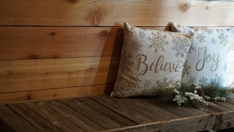 Two decorative pillows rest on a wooden bench. The pillows feature winter-themed design elements, including snowflakes, with the words 'Believe' and 'Joy' written on them. Pinecone and evergreen decorations are placed in front of the pillows, adding a festive touch. The wooden backdrop and bench contribute to a rustic ambiance.