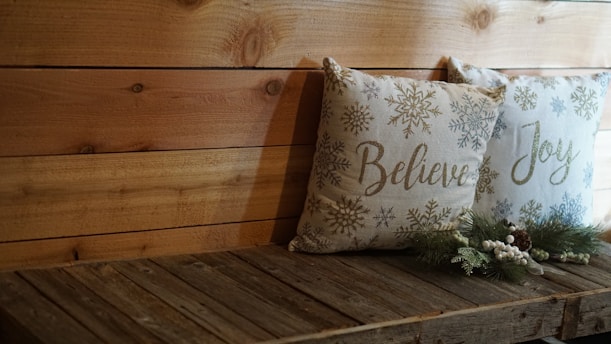 Two decorative pillows rest on a wooden bench. The pillows feature winter-themed design elements, including snowflakes, with the words 'Believe' and 'Joy' written on them. Pinecone and evergreen decorations are placed in front of the pillows, adding a festive touch. The wooden backdrop and bench contribute to a rustic ambiance.