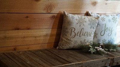 Two decorative pillows rest on a wooden bench. The pillows feature winter-themed design elements, including snowflakes, with the words 'Believe' and 'Joy' written on them. Pinecone and evergreen decorations are placed in front of the pillows, adding a festive touch. The wooden backdrop and bench contribute to a rustic ambiance.