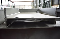 A close-up view of a large, metallic, industrial I-beam with visible welding marks. The image is taken inside a factory setting, with blurred background details indicating a spacious industrial interior. The beam has two large circular holes and appears rough and unfinished.