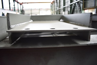 A close-up view of a large, metallic, industrial I-beam with visible welding marks. The image is taken inside a factory setting, with blurred background details indicating a spacious industrial interior. The beam has two large circular holes and appears rough and unfinished.