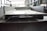 A close-up view of a large, metallic, industrial I-beam with visible welding marks. The image is taken inside a factory setting, with blurred background details indicating a spacious industrial interior. The beam has two large circular holes and appears rough and unfinished.