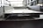 A close-up view of a large, metallic, industrial I-beam with visible welding marks. The image is taken inside a factory setting, with blurred background details indicating a spacious industrial interior. The beam has two large circular holes and appears rough and unfinished.