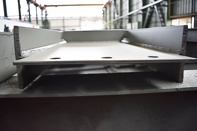 A close-up view of a large, metallic, industrial I-beam with visible welding marks. The image is taken inside a factory setting, with blurred background details indicating a spacious industrial interior. The beam has two large circular holes and appears rough and unfinished.