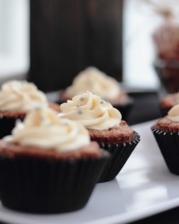 Several cupcakes with creamy, swirled frosting topped with small silver sprinkles are arranged on a white plate. The cupcakes are placed in dark-colored wrappers, and the soft focus background hints at a cozy, inviting setting.