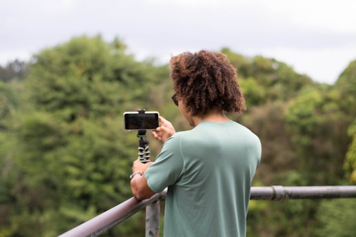 A person with curly hair is standing on a balcony or platform, holding a smartphone mounted on a flexible tripod while surrounded by lush greenery. The individual is focused on recording or capturing the scenery.