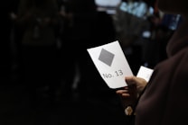 A hand holds a card labeled 'No. 13' featuring a gray diamond shape. The background is dark, with indistinct figures and objects.