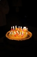 A birthday cake topped with colorful, lit candles arranged to spell 'Happy Birthday' is set against a dark background. The warm glow from the candles illuminates the cake, creating a cozy and celebratory atmosphere.