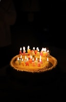 A birthday cake topped with colorful, lit candles arranged to spell 'Happy Birthday' is set against a dark background. The warm glow from the candles illuminates the cake, creating a cozy and celebratory atmosphere.
