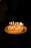 A birthday cake topped with colorful, lit candles arranged to spell 'Happy Birthday' is set against a dark background. The warm glow from the candles illuminates the cake, creating a cozy and celebratory atmosphere.