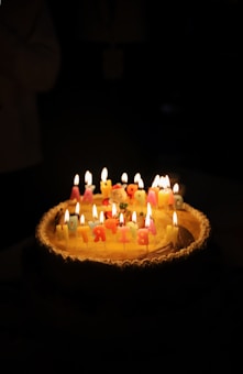 A birthday cake topped with colorful, lit candles arranged to spell 'Happy Birthday' is set against a dark background. The warm glow from the candles illuminates the cake, creating a cozy and celebratory atmosphere.