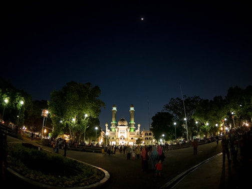 A night scene featuring a brightly lit mosque with multiple towers and a large dome, surrounded by lush green trees. The sky is dark, with a small crescent moon visible. A large crowd of people is gathered in the plaza in front of the mosque, illuminated by streetlights.