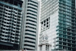 An urban landscape featuring modern high-rise buildings with glass facades, alongside an ornate clock tower. The architecture blends contemporary and classical elements, highlighting the contrast between sleek, reflective surfaces and intricate, historical design.