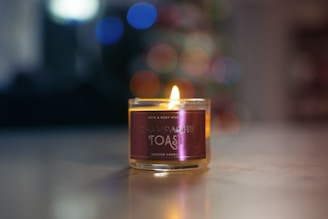 A lit scented candle labeled 'Champagne Toast' is placed on a reflective surface. The background features blurred, colorful lights creating a bokeh effect, suggesting a warm and cozy atmosphere.