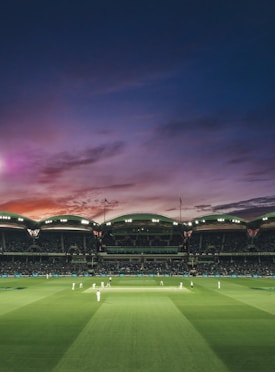 A cricket match taking place in a large stadium under a beautifully colored evening sky. The field is well-lit and surrounded by spectators, with players dressed in white uniforms actively engaged in the game.