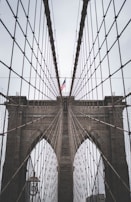 A close-up perspective of a suspension bridge showcasing its intricate cable design and towering stone arches. An American flag is prominently displayed at the center.