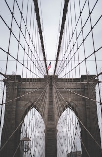 A close-up perspective of a suspension bridge showcasing its intricate cable design and towering stone arches. An American flag is prominently displayed at the center.