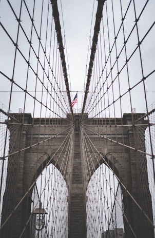 A close-up perspective of a suspension bridge showcasing its intricate cable design and towering stone arches. An American flag is prominently displayed at the center.