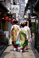 A narrow alley lined with traditional Japanese shops and signs, with three women dressed in traditional kimonos walking away from the camera. The women have intricate hairstyles adorned with hair accessories and ornate sashes on their kimonos. The alley is decorated with lanterns and various signboards, and appears to be in an older part of a city.