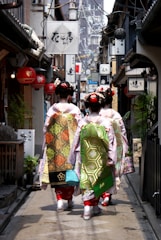 A narrow alley lined with traditional Japanese shops and signs, with three women dressed in traditional kimonos walking away from the camera. The women have intricate hairstyles adorned with hair accessories and ornate sashes on their kimonos. The alley is decorated with lanterns and various signboards, and appears to be in an older part of a city.