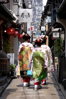 A narrow alley lined with traditional Japanese shops and signs, with three women dressed in traditional kimonos walking away from the camera. The women have intricate hairstyles adorned with hair accessories and ornate sashes on their kimonos. The alley is decorated with lanterns and various signboards, and appears to be in an older part of a city.