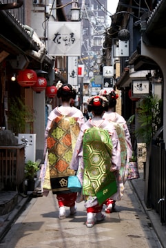A narrow alley lined with traditional Japanese shops and signs, with three women dressed in traditional kimonos walking away from the camera. The women have intricate hairstyles adorned with hair accessories and ornate sashes on their kimonos. The alley is decorated with lanterns and various signboards, and appears to be in an older part of a city.
