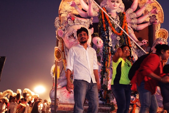 A group of people are gathered in front of a large, ornate statue of a multi-armed deity. It is decorated with garlands and flowers, and the scene is set during the evening. The atmosphere is lively with people moving around, possibly at a festival or religious celebration.