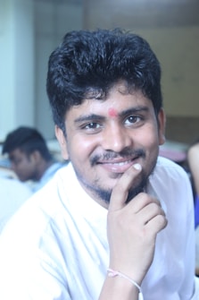 A person with short, curly hair and a friendly smile. They have a small red mark on their forehead and are resting their hand on their chin. The background appears to be indoors, possibly a classroom or office, with another person slightly out of focus.