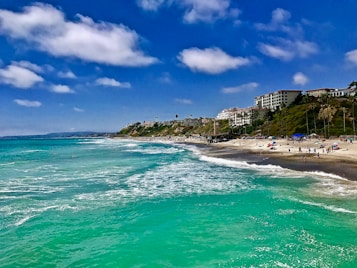 A picturesque coastline with turquoise water gently lapping on a sandy beach. In the background, white buildings and palm trees line the hillside, with a few people scattered along the shoreline enjoying the sunny weather. The sky is a vibrant blue with fluffy white clouds.