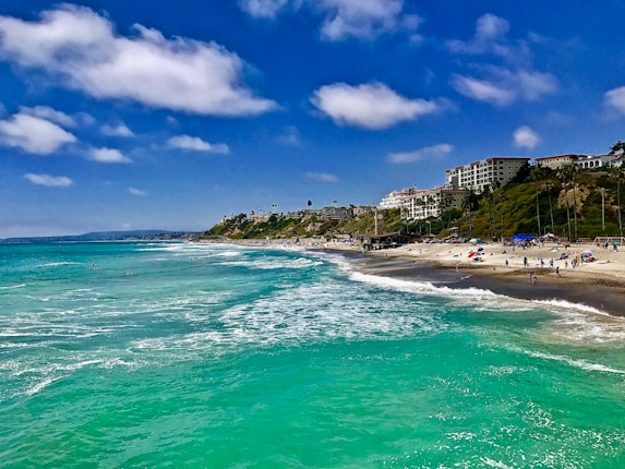 A picturesque coastline with turquoise water gently lapping on a sandy beach. In the background, white buildings and palm trees line the hillside, with a few people scattered along the shoreline enjoying the sunny weather. The sky is a vibrant blue with fluffy white clouds.